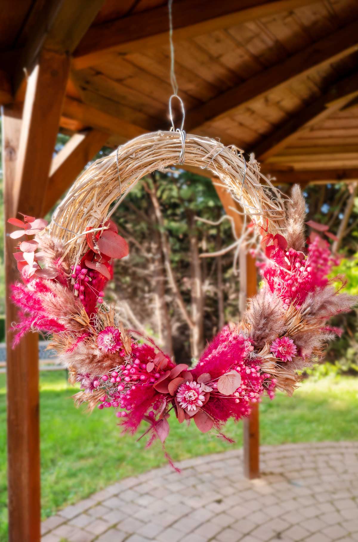 Rustikaler 'Naturliebe' Trockenblumenkranz an Holztür, handgemachte Wanddeko in lebhaften Rot- und Rosatönen, ideal für ein warmes Willkommen.